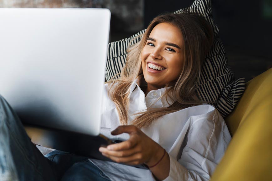 Frau liegt glücklich auf dem Sofa mit einem Laptop in den Händen.