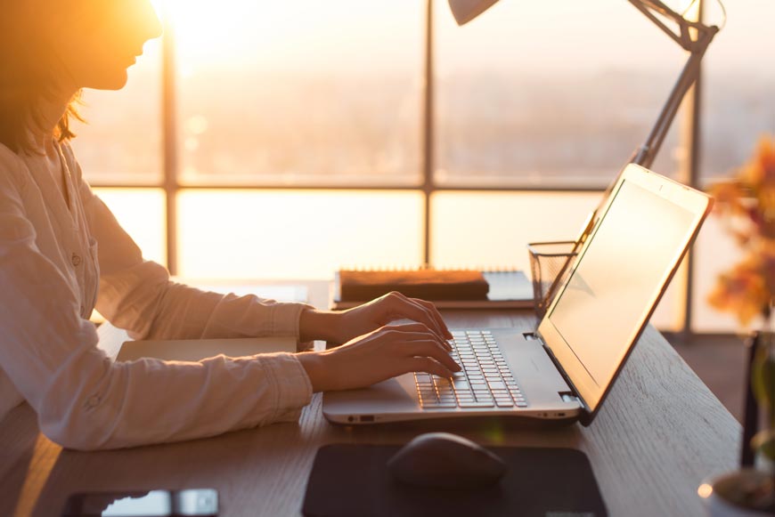 Dame sitzt vor großer Fensterfront bei Sonnenuntergang am Schreibtisch und arbeitet mit dem Laptop.